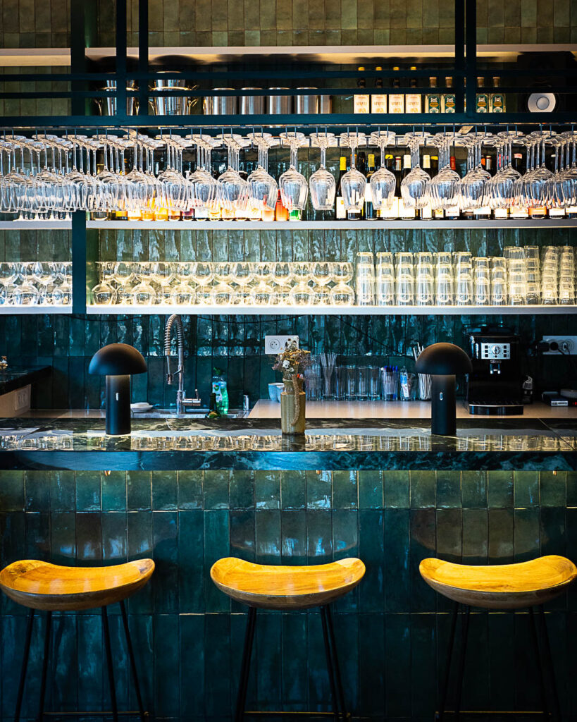 Photo du bar à vin Euphorie. Sur celle-ci on voit le bar du restaurant avec ses carreaux verts foncés, ses dizaines de verres à vin suspendus et ses tabourets jaunes.