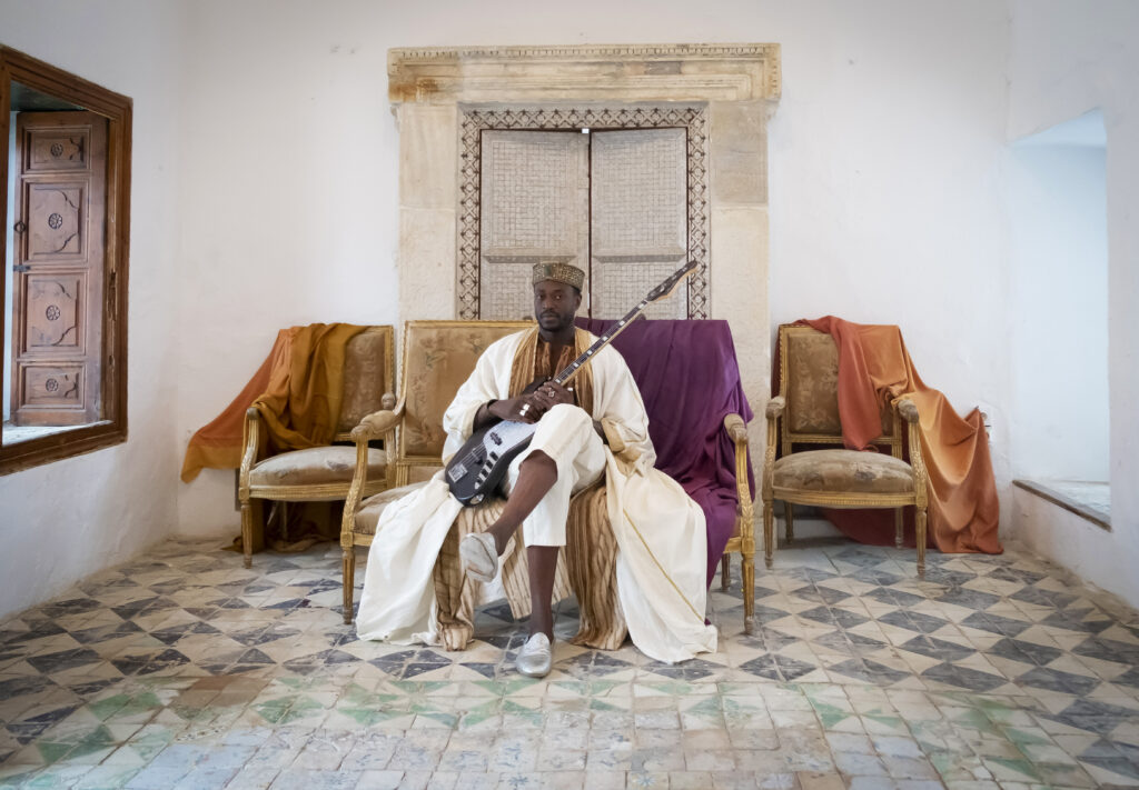 Photo Alune Wade assis sur un fauteuil en tenue traditionnelle sénégalaise avec sa basse à la main