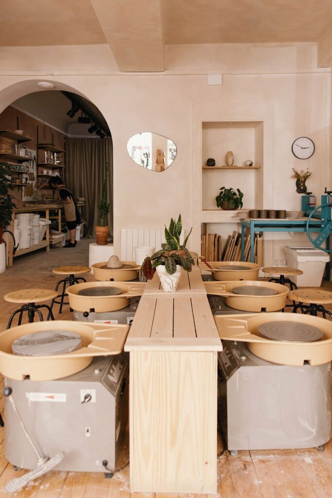 Photo de l'atelier de poterie avec des plaques tournantes, de l'argile, et des tabourets. L'ambiance est lumineuse et chaleureuse 