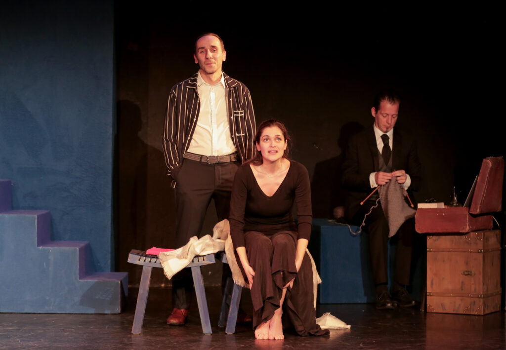 Photo d'une scène issue de la pièce de théâtre avec une femme assise un homme debout derrière elle et un autre homme tricotant derrière elle.